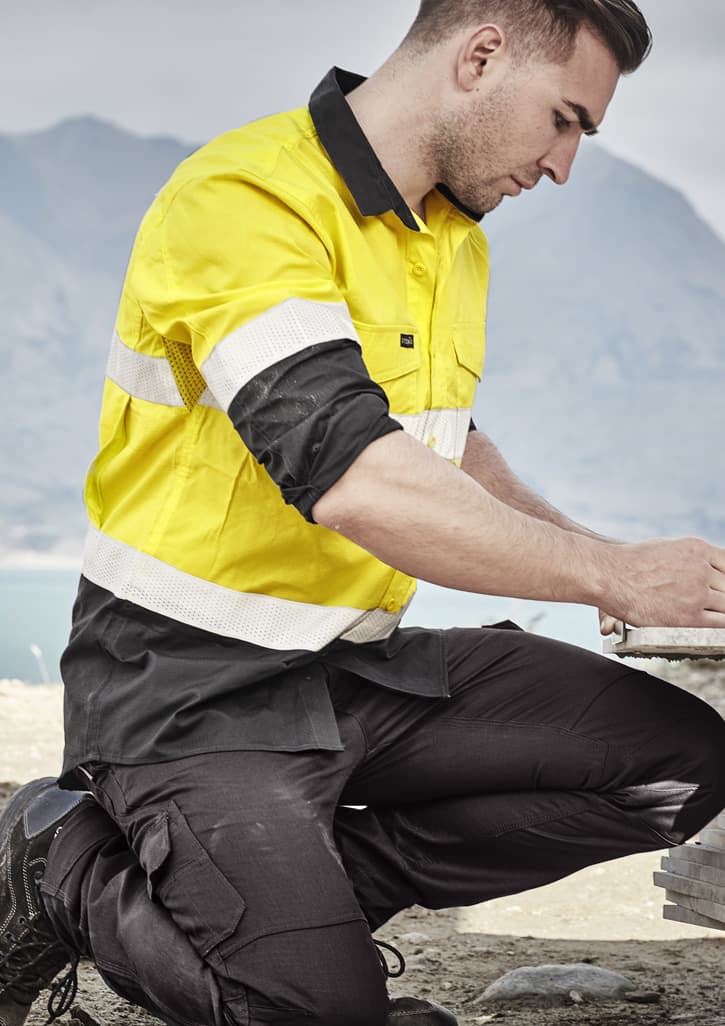 A man kneeling outdoors in a yellow and black long sleeve work shirt with reflective tape, engaged in construction work.