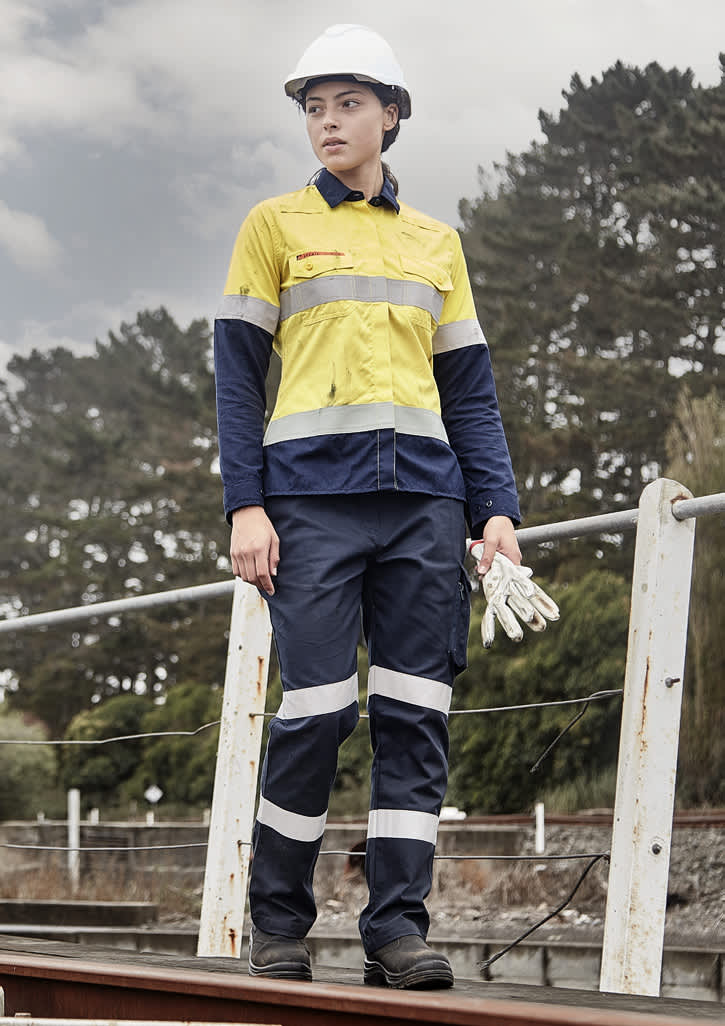 A woman in a yellow and navy work shirt with reflective stripes, wearing a hard hat and gloves, stands outdoors.