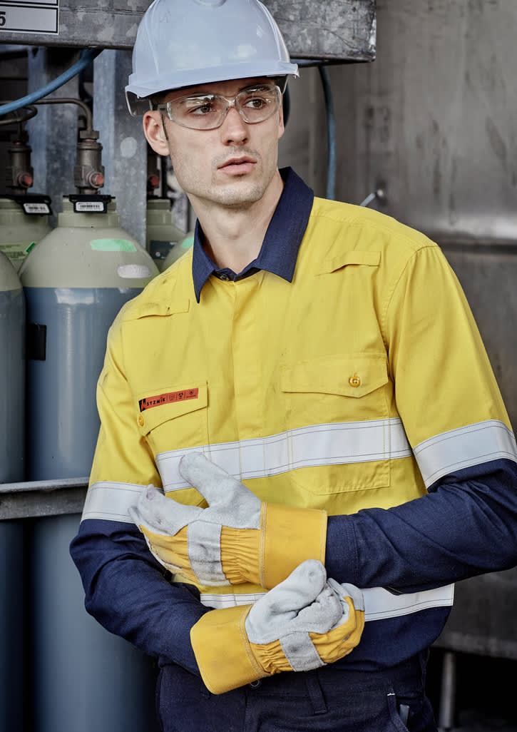 A man in a yellow and navy work shirt with reflective stripes, wearing protective goggles and gloves, standing near gas cylinders.