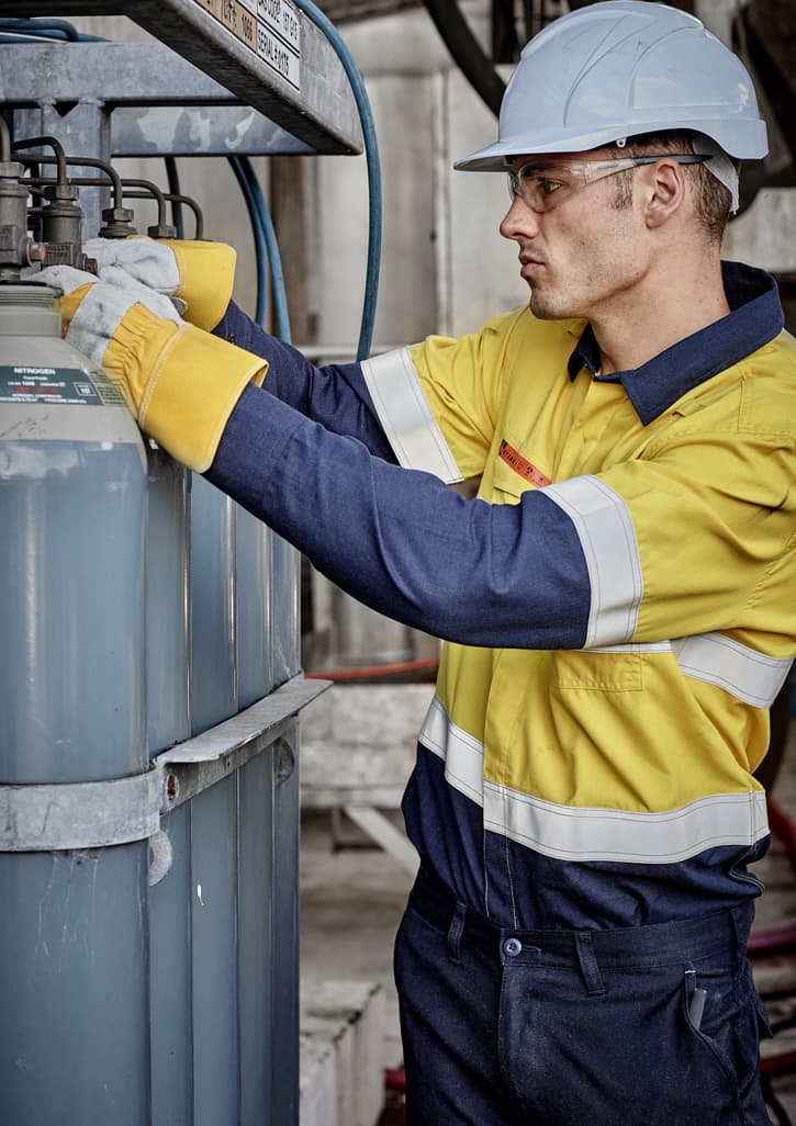A worker in a yellow and navy spliced shirt adjusts equipment, wearing a hard hat and gloves in an industrial setting.