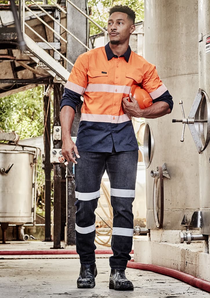 A worker in an orange and navy long sleeve shirt with segmented tape, holding a helmet in an industrial setting.