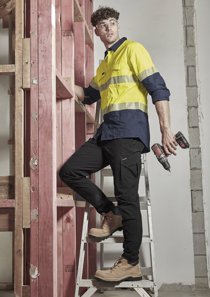 A man in a yellow and navy long sleeve shirt stands on a ladder, holding power tools, next to timber framing.