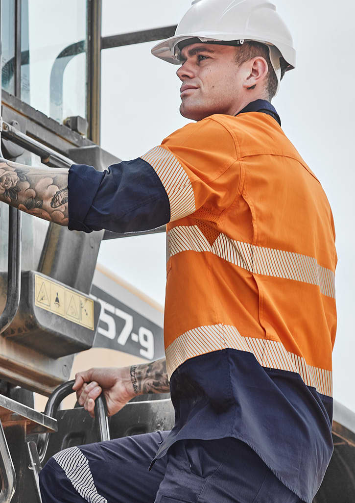 A man in an orange and navy long sleeve hi-vis shirt with segmented tape climbs a machine, wearing a white hard hat.