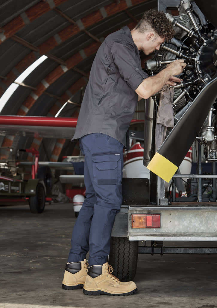 A man in a dark grey shirt and navy pants works on a machine, wearing tan boots in a hangar setting.