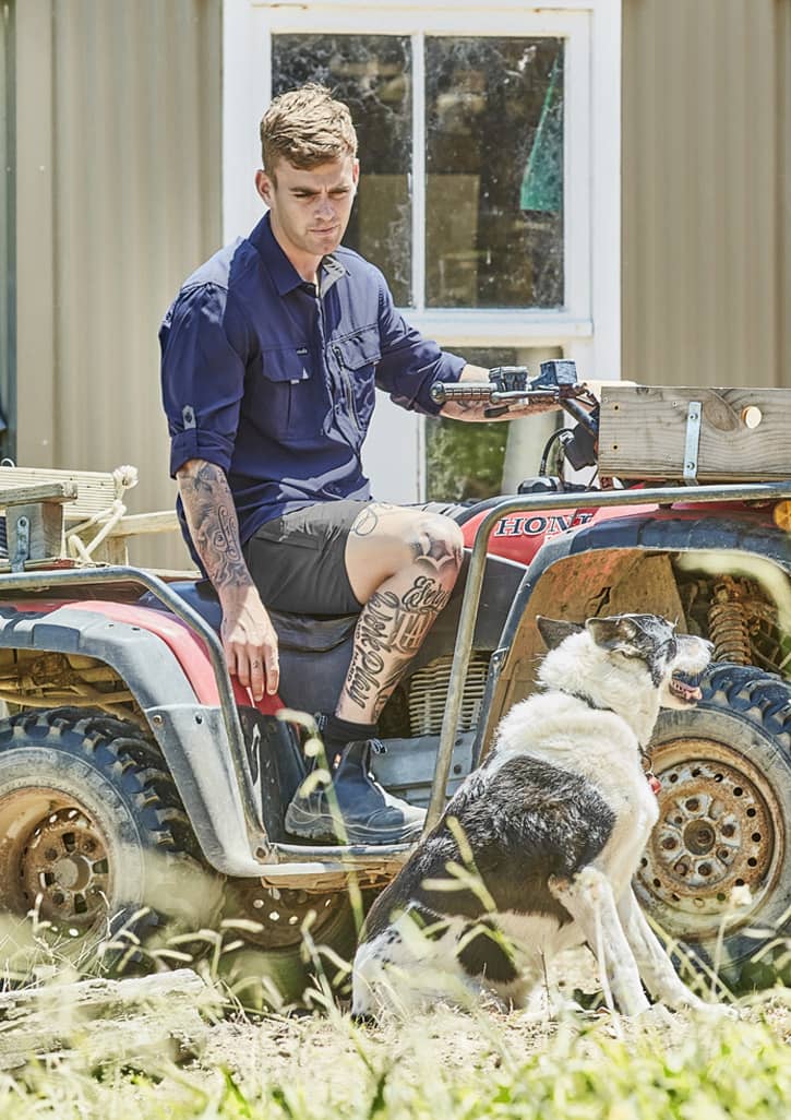 A man in a navy long sleeve button-up shirt sits on an all-terrain vehicle beside a dog. The setting is outdoor with grass.