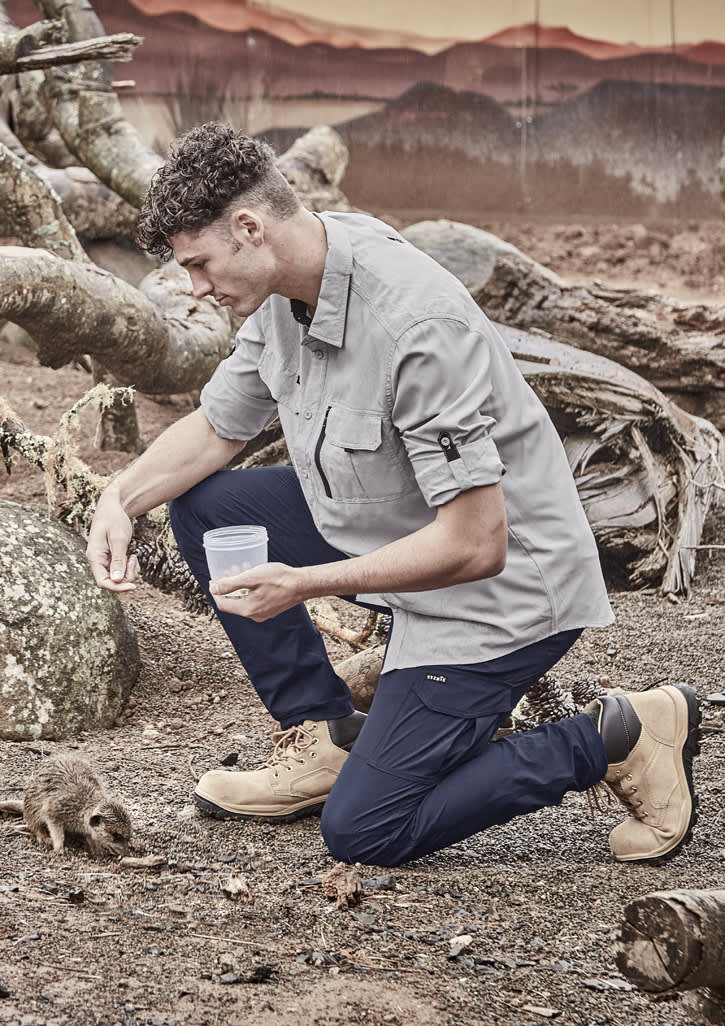 A man kneels in a grey long-sleeve button-up shirt and navy pants, interacting with a small animal outdoors.