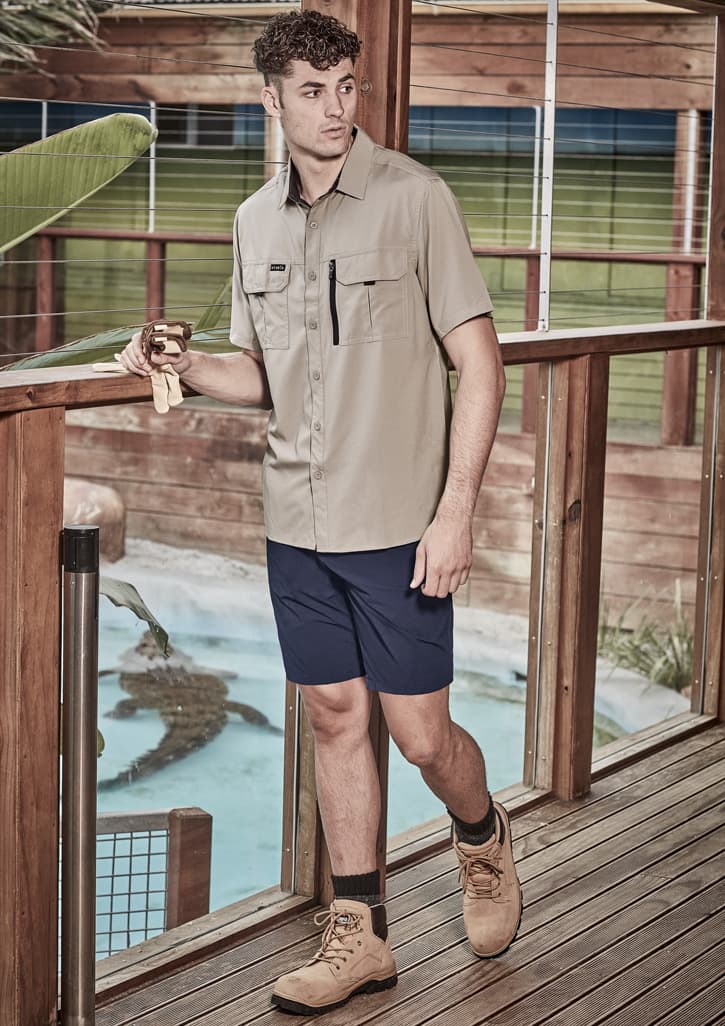 A man wearing a light brown short sleeve button-up shirt and navy shorts, standing on a wooden deck.
