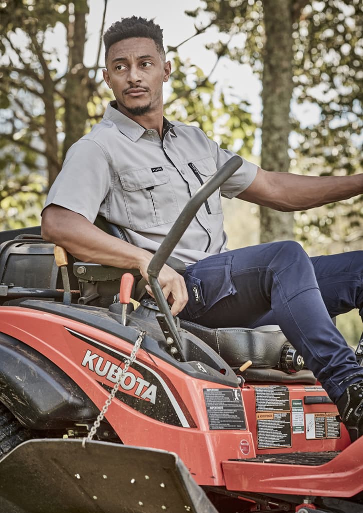 A man wearing a grey short sleeve button-up shirt sits on a red Kubota lawn mower amidst greenery.