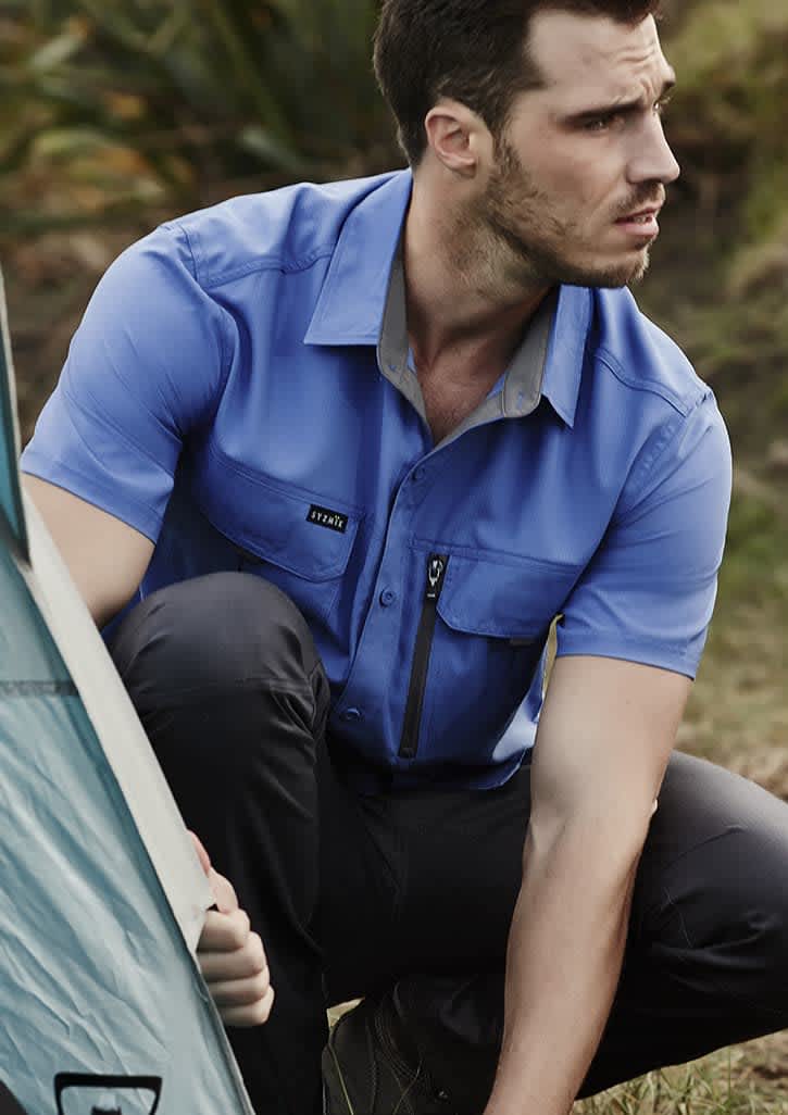 A blue short sleeve button-up shirt with a chest pocket and a logo, worn by a man outdoors.