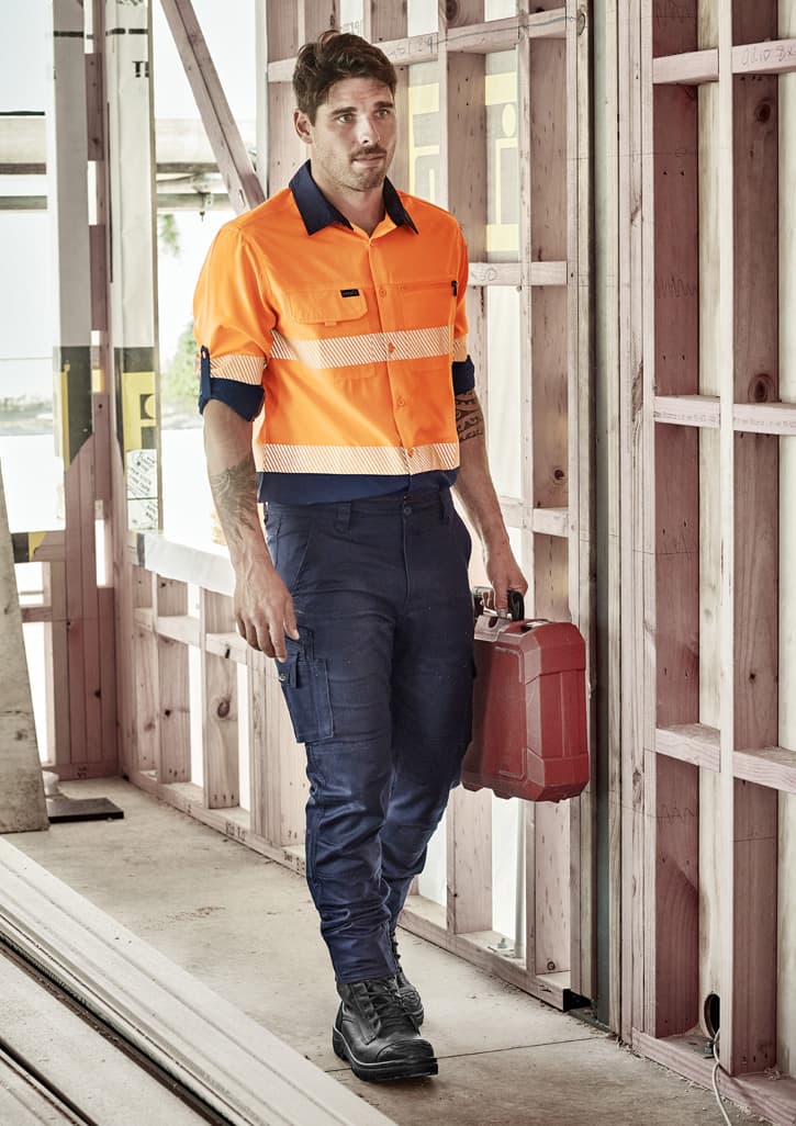A man wearing an orange long sleeve segmented tape work shirt and navy pants, holding a red toolbox in a construction setting.