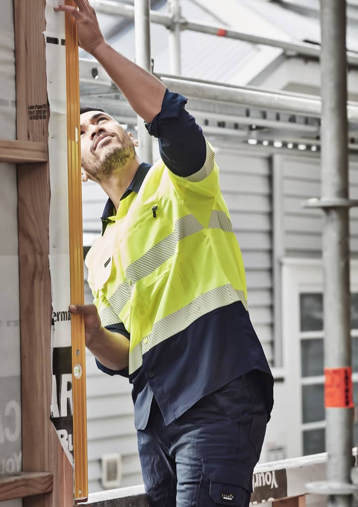 A man wearing a hi-vis yellow and navy long sleeve work shirt examines a construction site using a level tool.