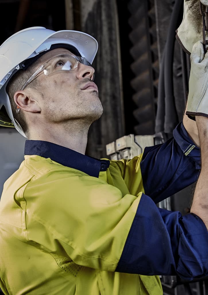A man in a yellow and navy long sleeve shirt with a helmet and safety glasses, examining equipment.