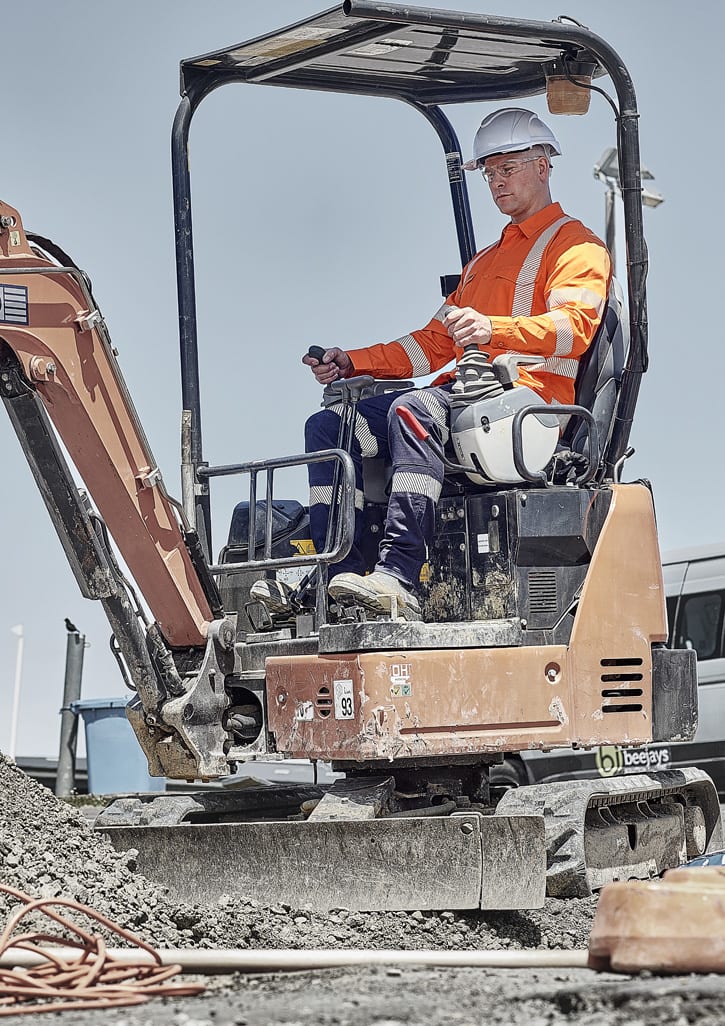A worker in a high-visibility orange shirt and white hard hat operates a small excavator on a construction site.