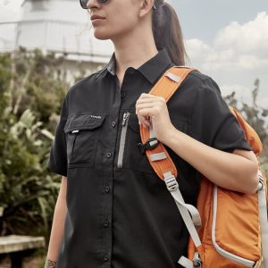 A woman in a black short sleeve outdoor shirt carries an orange backpack, standing near a lighthouse.