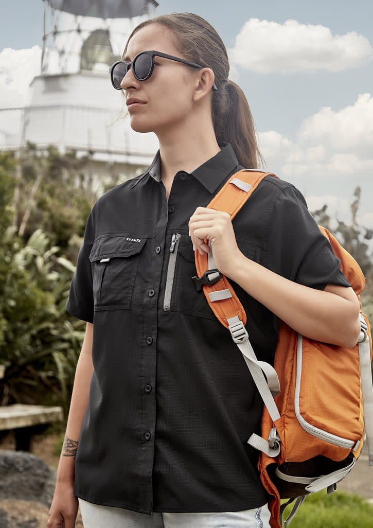 A woman in a black short sleeve outdoor shirt carries an orange backpack, standing near a lighthouse.