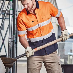 A short sleeve work shirt in orange and navy with reflective tape, worn by a man using a shovel, with a construction setting in the background.
