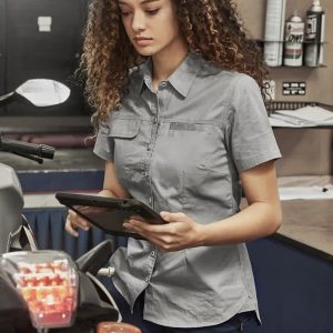 Lightweight grey short sleeve shirt for women, featuring a front button closure and a logo, displayed in a workshop setting.