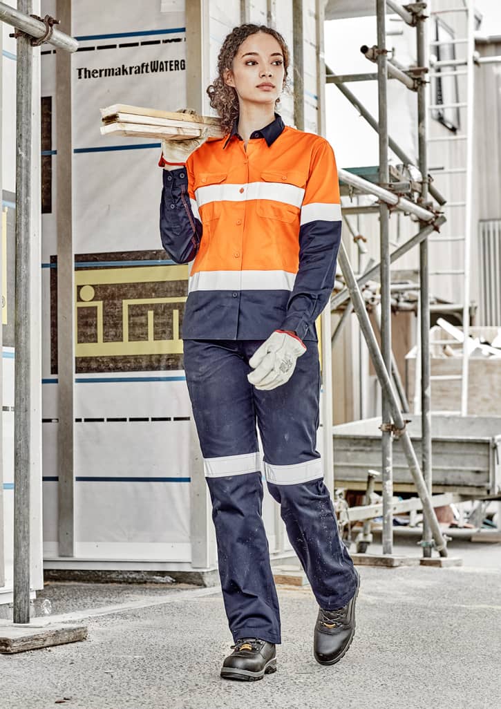 A woman in an orange and navy long sleeve industrial shirt, holding timber, wears work gloves and sturdy boots.