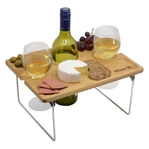 A wooden picnic table with two glasses of white wine, a bottle, cheese, crackers, olives, and grapes.