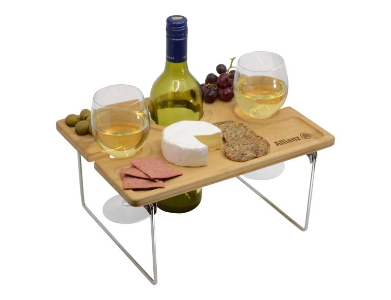 A wooden picnic table with two glasses of white wine, a bottle, cheese, crackers, olives, and grapes.