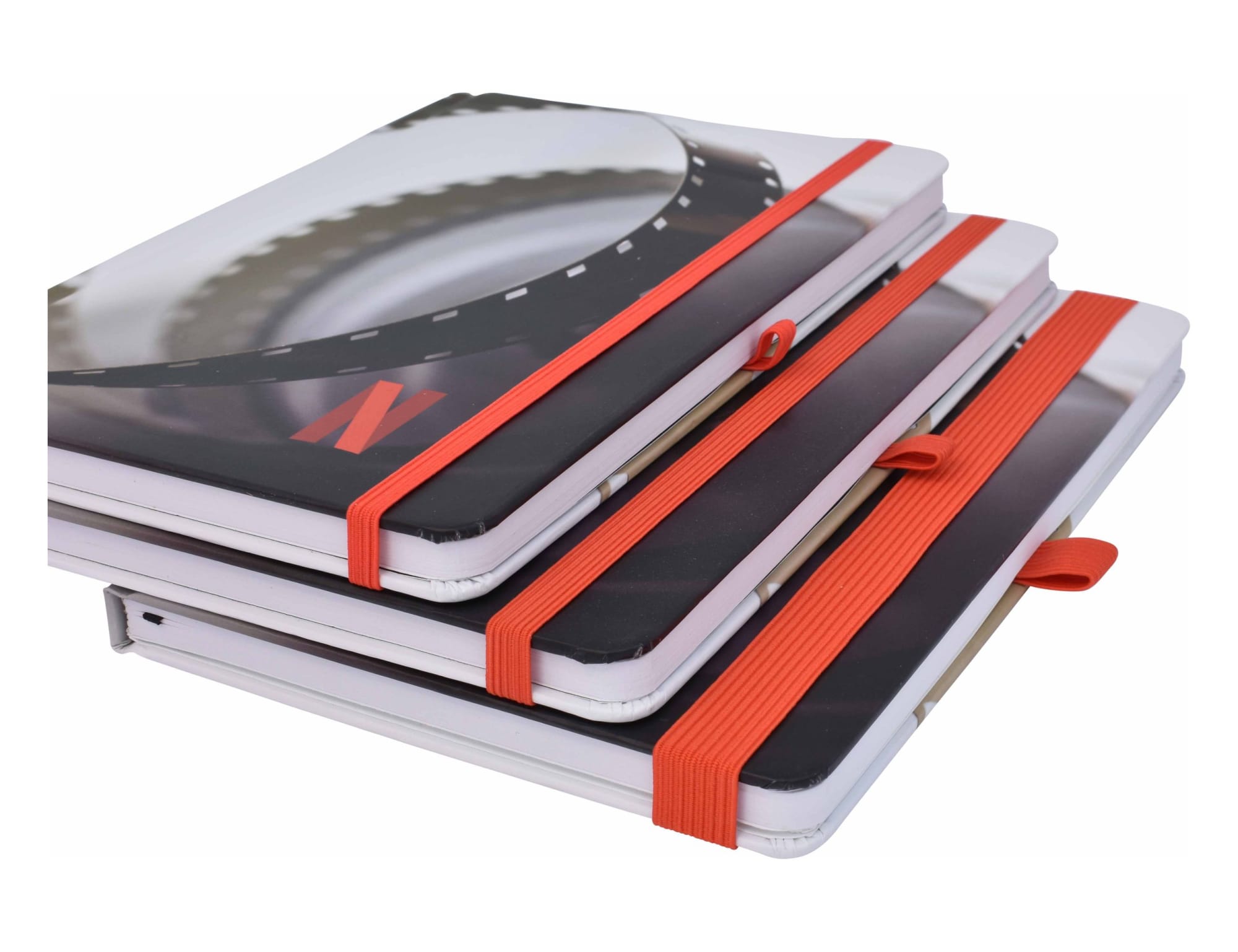A set of three A5 notebooks featuring full-colour designs and orange elastic bands. The notebooks have a sleek, matte finish.