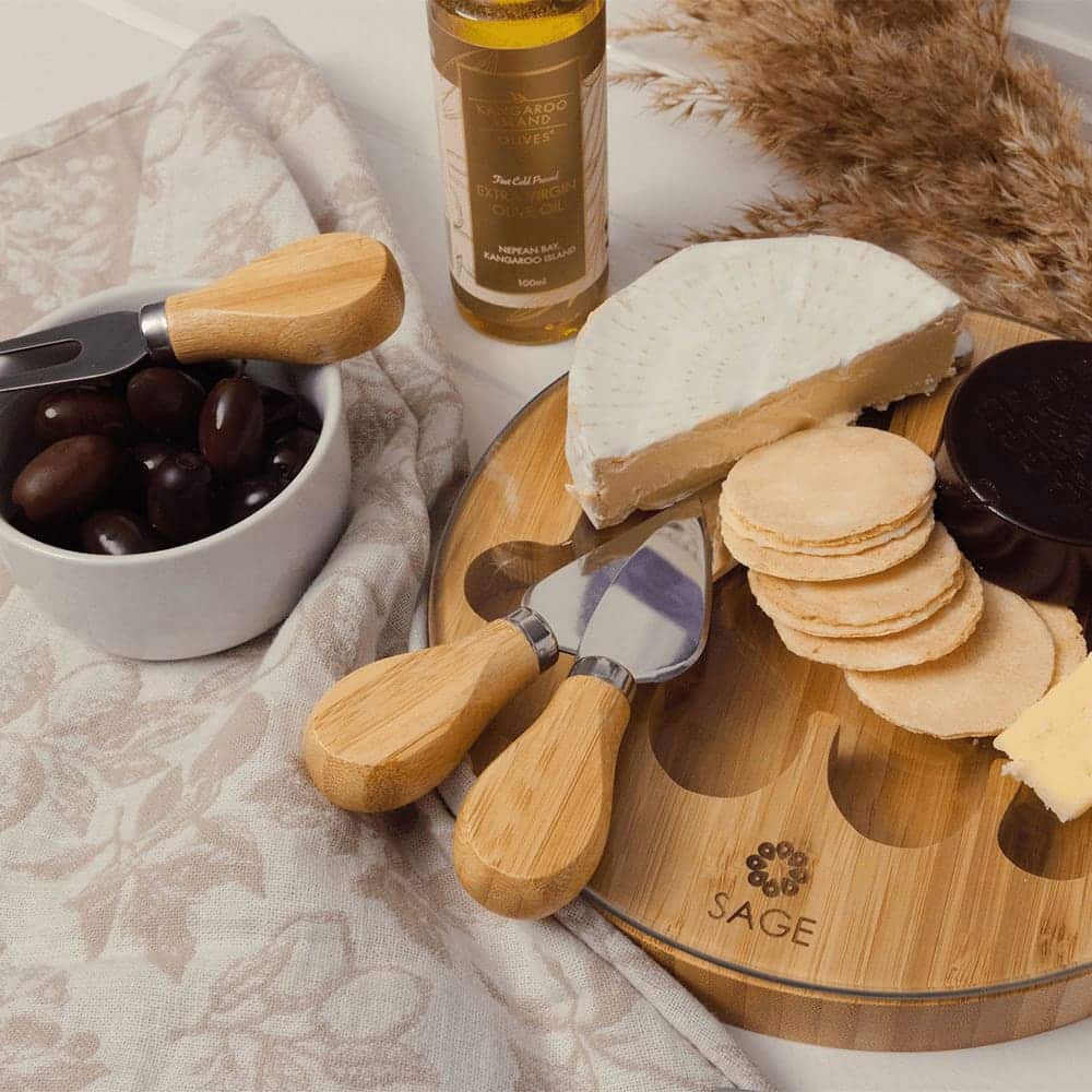 A wooden cheese board with a round cheese, crackers, and a bowl of olives, accompanied by cheese knives.