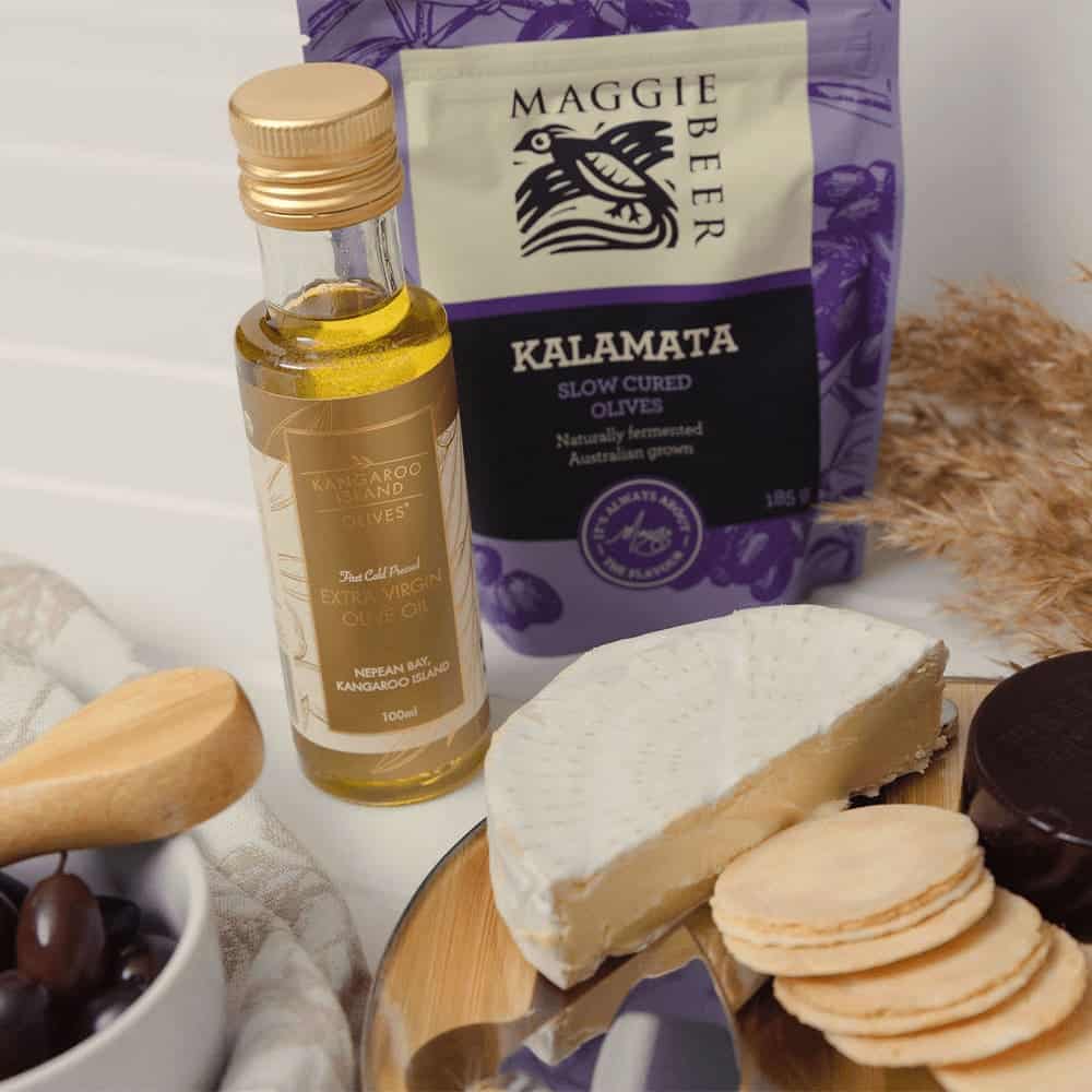 A gourmet gift pack with cheese, olives, olive oil, and biscuits, featuring a purple bag and wooden serving utensils.
