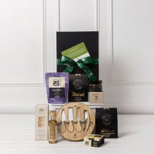 A gourmet gift pack featuring a wooden serving board, assorted gourmet snacks, olive oil, and a jar of jam, all in a black box.
