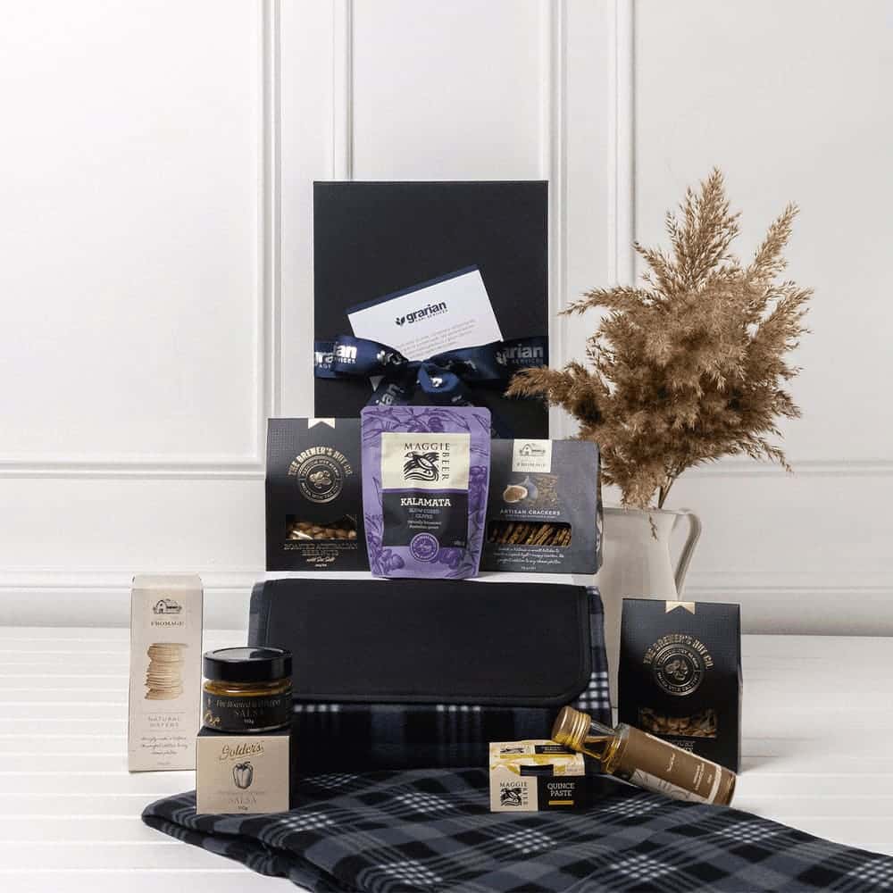 A stylish black gift box containing gourmet snacks, a picnic blanket, and packaged treats, featuring a decorative arrangement.