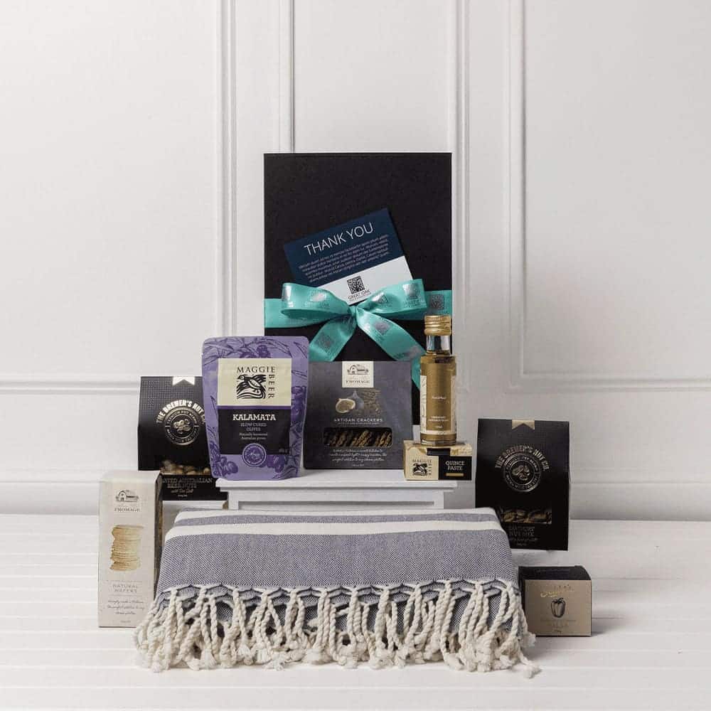 A gourmet gift pack featuring a grey and white throw, assorted treats, and a small bottle, all in elegant packaging.