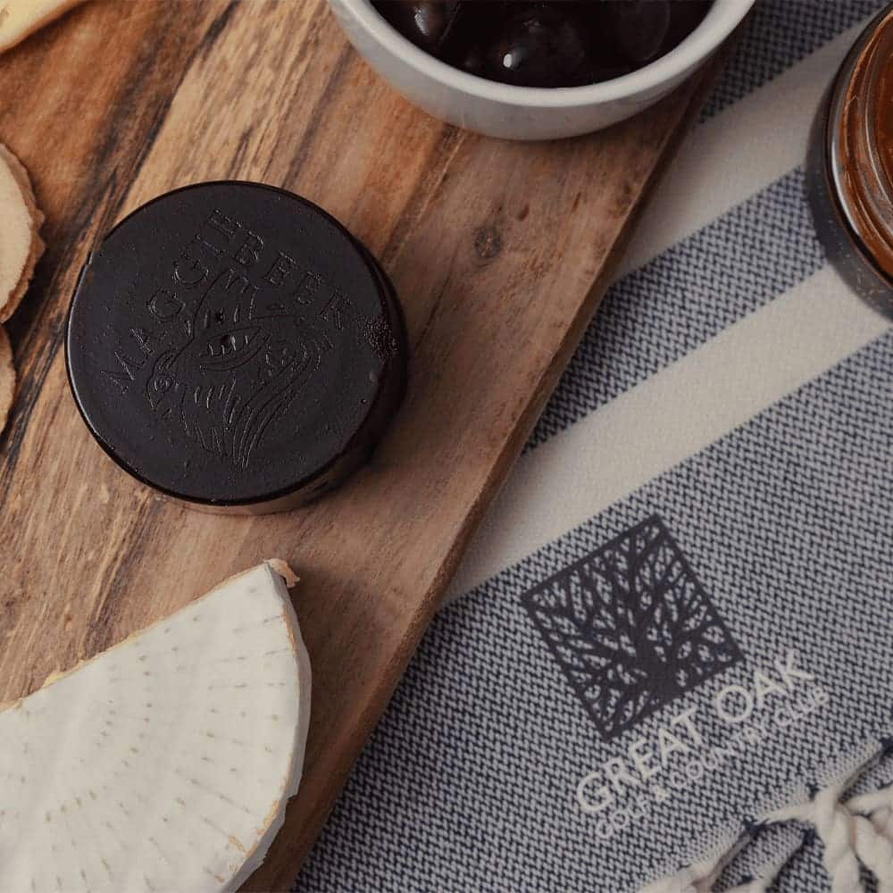 A wooden platter featuring a round black cheese, olives, and a slice of cheese, alongside a textured cloth with branding.