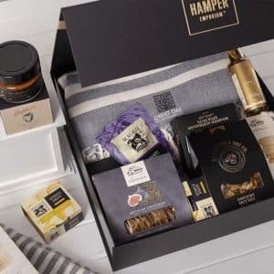 A gift pack featuring a grey blanket, gourmet snacks, nut mix, crackers, and a jar of preserve, all in a black box.