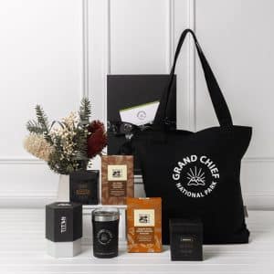 A black tote bag with a logo, surrounded by various sweet treat boxes, a candle, and decorative dried flowers.