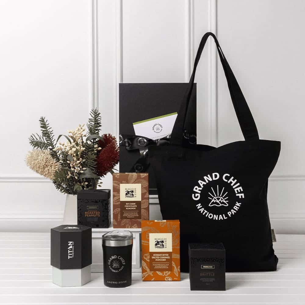 A black tote bag with a logo, surrounded by various sweet treat boxes, a candle, and decorative dried flowers.