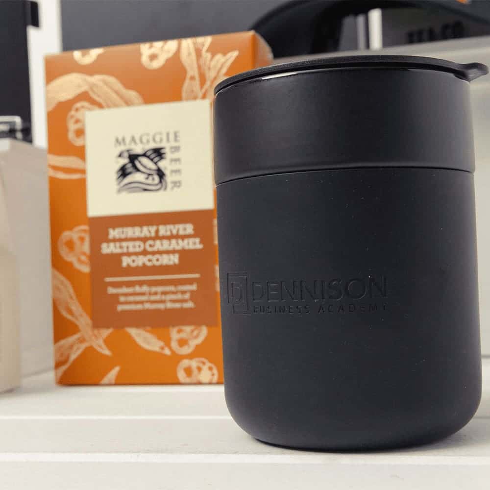 A black insulated drink container with a lid beside a box of salted caramel popcorn, featuring a logo on the container.