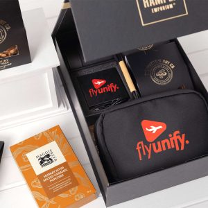 A black gift box contains a branded tech kit, featuring a black pouch, notebook, pen, and snacks.