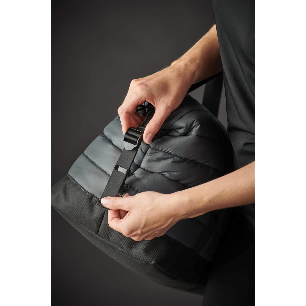 A person adjusting the strap of a black and grey quilted duffle bag, featuring a textured finish and a logo.
