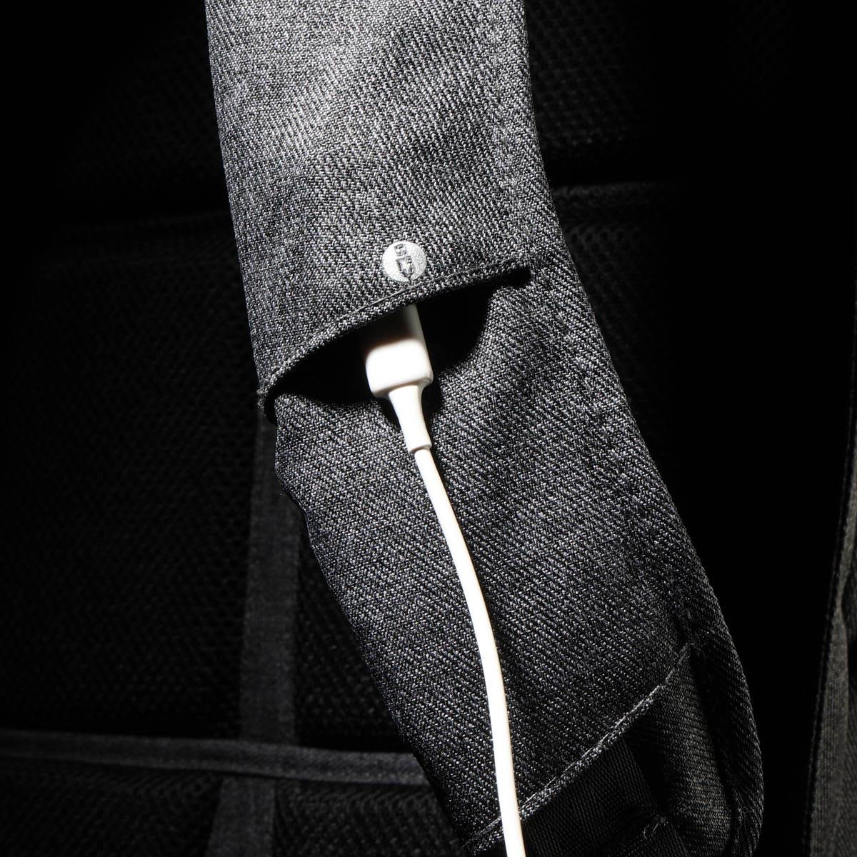 A close-up of a black backpack strap featuring a white charging cable tucked inside. The strap has a logo.