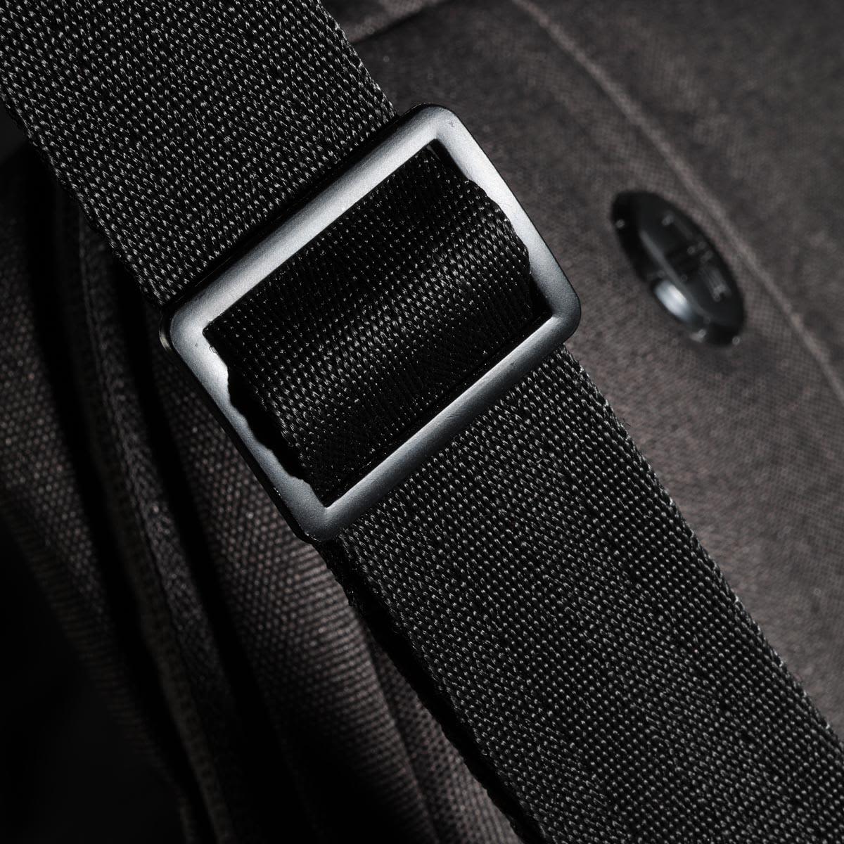 A close-up of a black strap buckle on a bag, featuring a textured fabric and a circular logo.