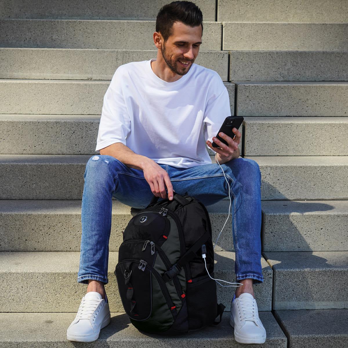 A man sits on steps, holding a phone, with a black Swissdigital Vector Backpack beside him.