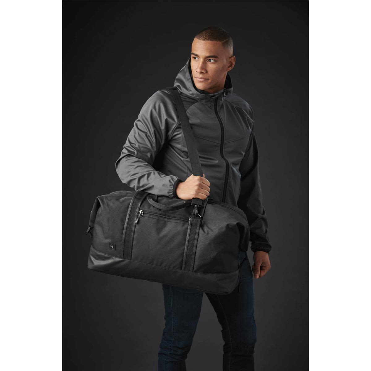 A man in a grey jacket carries a large black duffle bag featuring a logo.