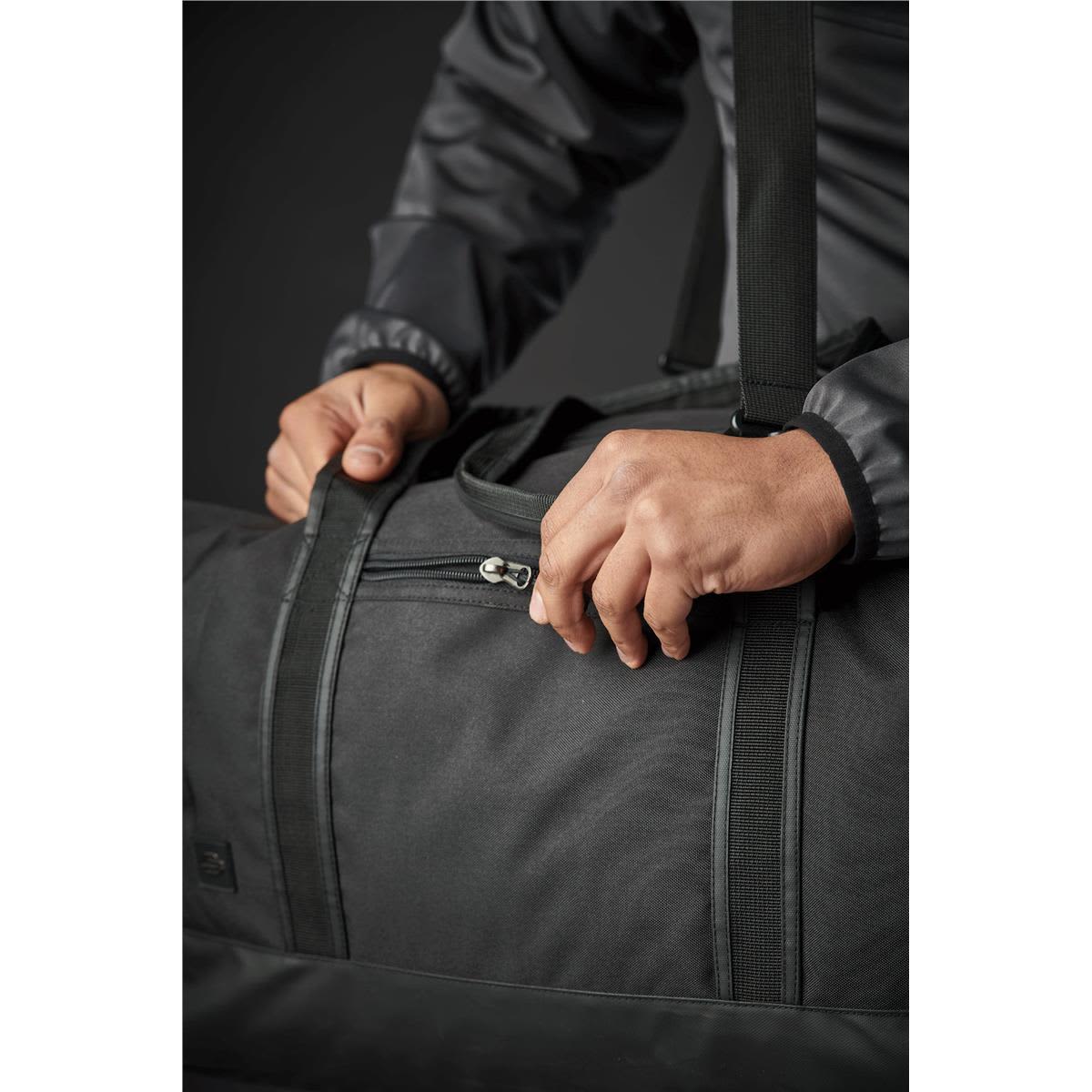 A dark green duffle bag with a zippered opening, being held by a person with a grey jacket. Features a logo.