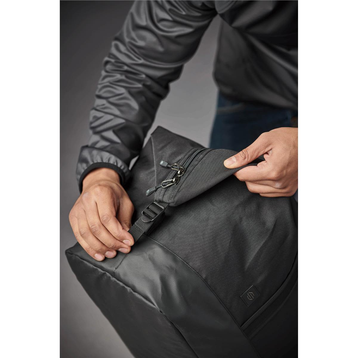 A person holding a black duffle bag, partially unzipping it, showcasing its sleek design and a logo.