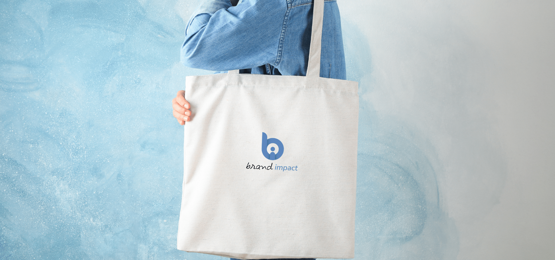 Person holding a natural cotton branded tote bag printed with the Brand Impact logo against a soft blue textured background.