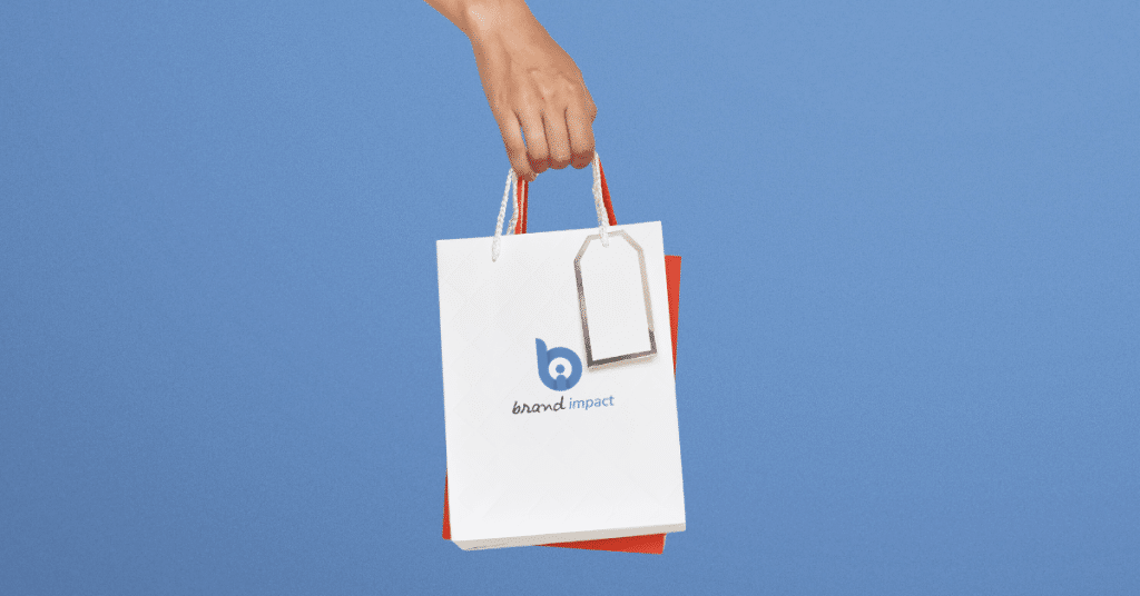 Hand holding a branded paper shopping bag with Brand Impact logo and blank gift tag, representing custom promotional packaging and corporate gifting.