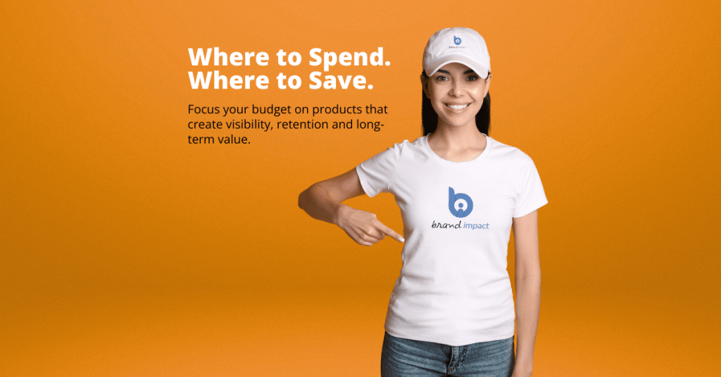 Woman wearing a white branded t-shirt and matching cap, pointing to a printed company logo on her shirt against an orange background. The banner features the headline “Where to Spend. Where to Save.” and promotes strategic promotional merchandise budgeting focused on visibility, customer retention and long-term brand value.