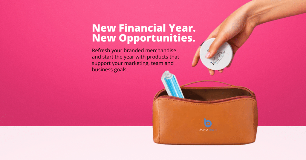Hand placing a branded compact mirror into a tan cosmetic bag with a branded logo against a pink background. The banner features the headline “New Financial Year. New Opportunities.” and encourages businesses to refresh their branded merchandise with practical promotional products that support marketing, team engagement and business goals.
