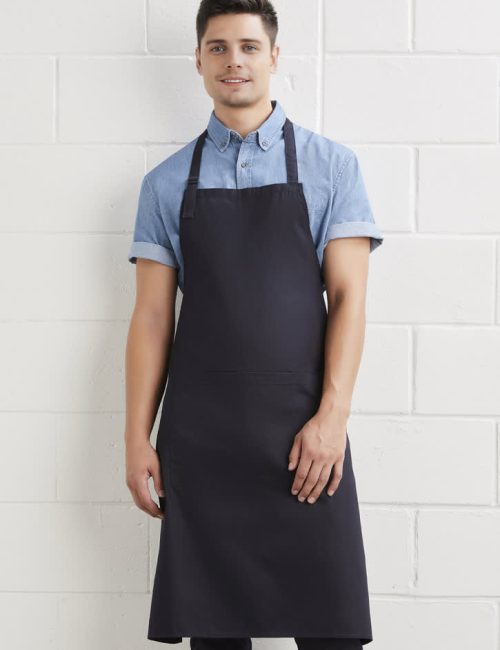 A man wearing a dark navy bib apron over a light blue shirt, standing against a white wall. The apron has a logo.