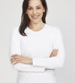 A white women's long sleeve performance tee, modelled by a smiling woman with arms crossed. The shirt has a logo.