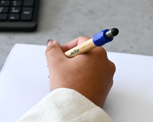 A bamboo spinner pen with a blue grip in a hand, resting on a lined notebook with a keyboard nearby.
